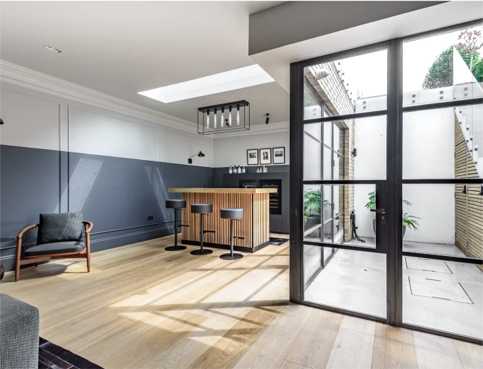 Modern basement conversion with a wooden bar, black stools, skylight, and large glass doors leading to an outdoor courtyard with a staircase.