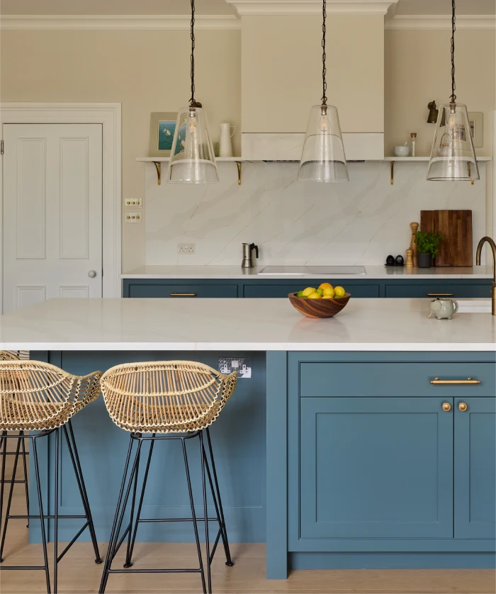 Elegant kitchen with blue cabinets, wicker bar stools, pendant lights, and a marble island topped with a fruit bowl