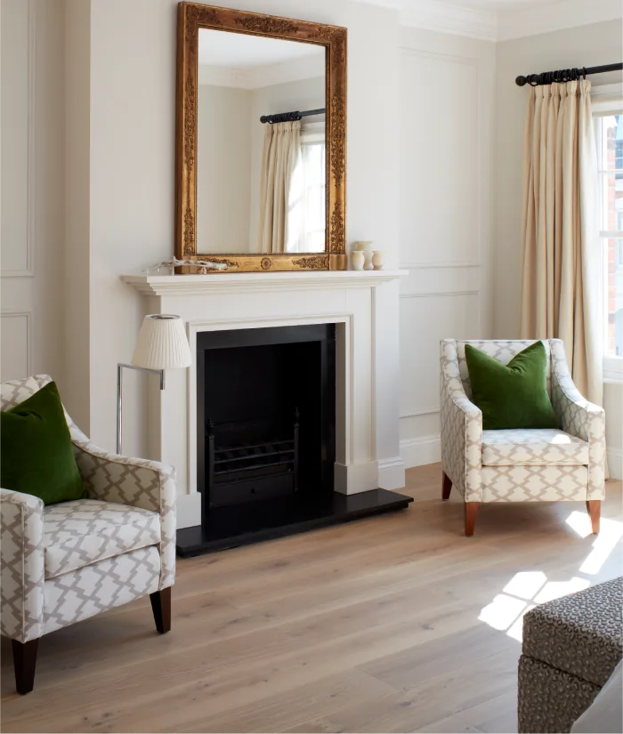 A cozy and elegant living room featuring a classic white fireplace with a large ornate gold mirror above it, flanked by patterned armchairs accented with green pillows, and natural light pouring through the cream curtains.