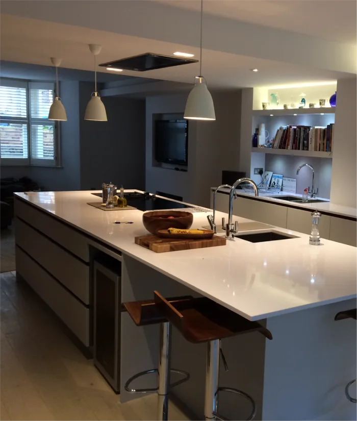 This modern kitchen boasts a sleek white countertop with a built-in sink and stove, complemented by elegant pendant lighting above. Open shelving with backlighting displays books and decorative items, while bar stools add functionality to the island.