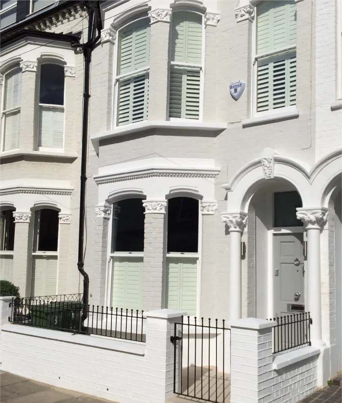 This charming white-painted facade features classic Victorian-style architecture, adorned with intricate moldings and arched details around the windows and door. The home includes shuttered windows and a small front yard enclosed by an elegant black wrought-iron fence.