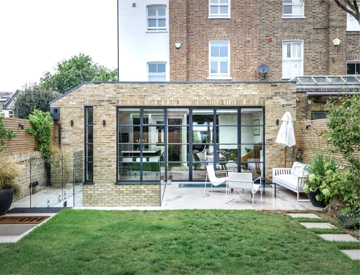 Modern house extension with brick walls, large glass doors, an outdoor patio area with seating, and a well-maintained green lawn.