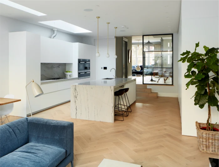 Modern open-plan kitchen with a marble island, pendant lighting, white cabinetry, herringbone wooden floors, and a cozy seating area with a navy sofa.