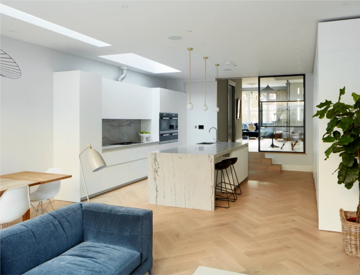 Bright modern kitchen extension with a marble island, skylights, herringbone flooring, and a blue sofa.