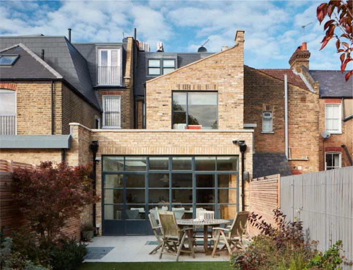 Contemporary home with a loft conversion, brick exterior, large glass doors, and a backyard featuring a wooden dining set and lush landscaping.