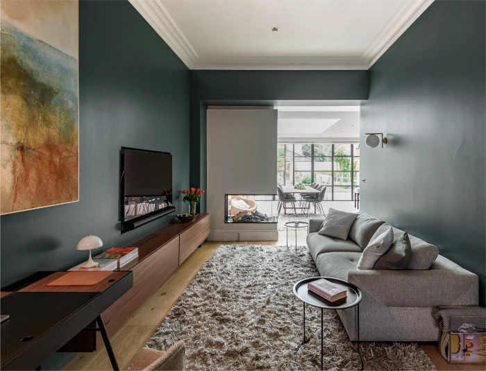 Elegant living room with a grey sofa, shaggy rug, mounted TV, wooden console, and a glass fireplace leading to a bright dining area with large windows