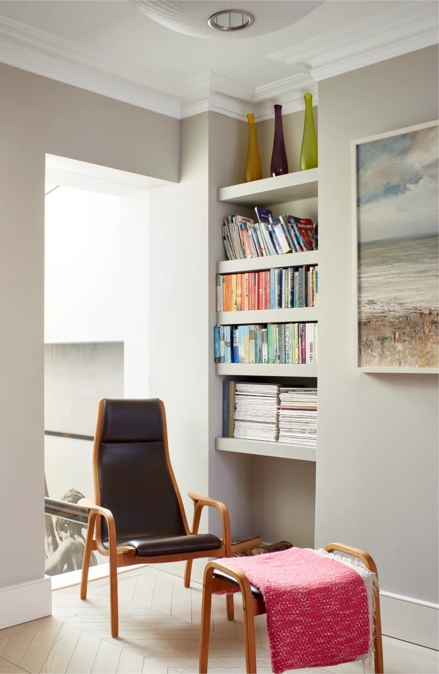 Cozy reading nook with a black leather chair, a small wooden table draped with a red blanket, built-in shelves filled with colorful books and magazines, and decorative vases.