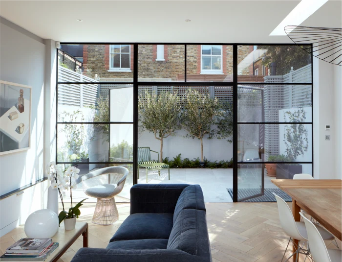 Bright modern living area with a navy sofa, glass coffee table, and floor-to-ceiling glass doors leading to a private courtyard with trees and patio seating.