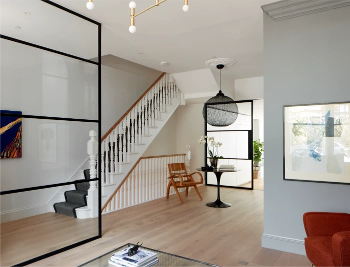 Modern interior with a white staircase, glass partitions, a round black pendant light, a wooden chair, and a framed artwork in a bright open space.