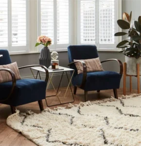 Chic living area with two navy armchairs, a geometric side table, a soft patterned rug, and a potted plant by a bay window with shutters.