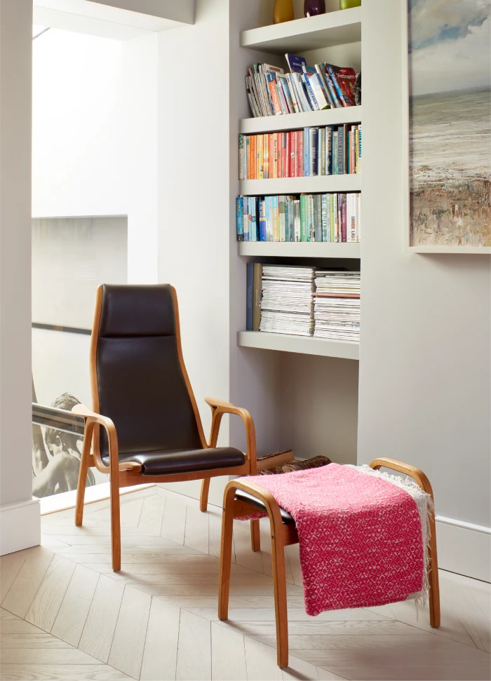 Cozy reading nook with a sleek black leather chair, matching wooden footrest draped in a red blanket, built-in shelves filled with colorful books and magazines.