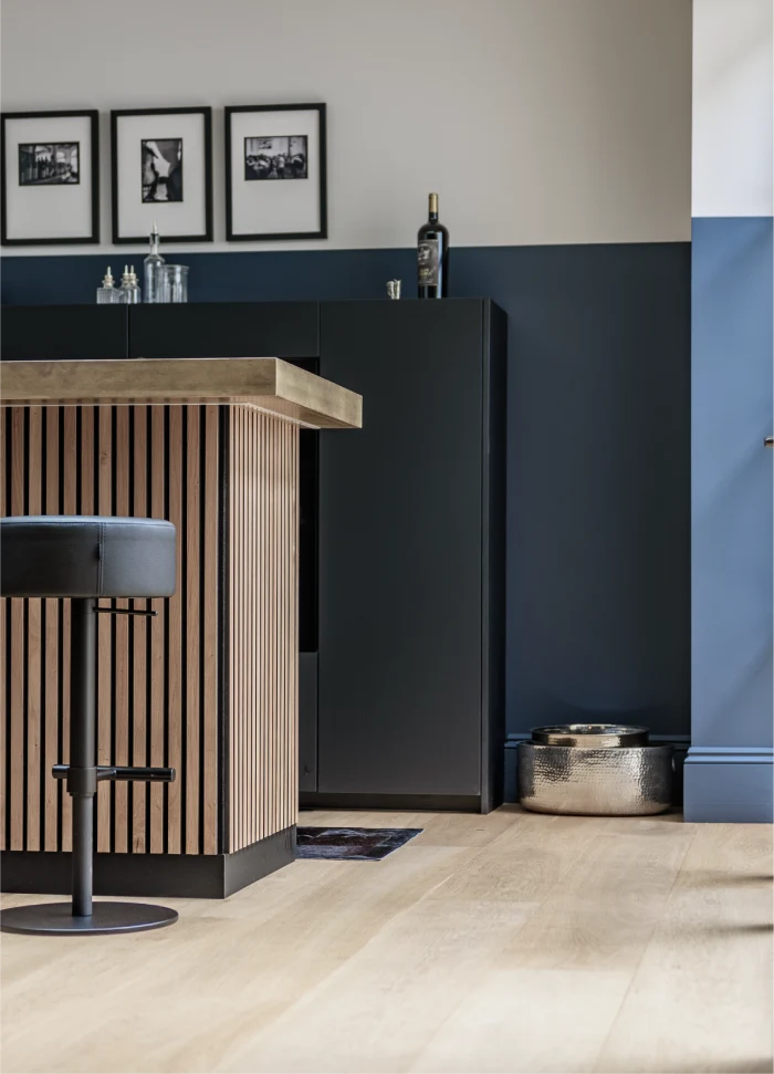 Close-up of a modern wooden kitchen island with vertical slats, a black bar stool, navy cabinetry, and framed black-and-white photos on the wall.