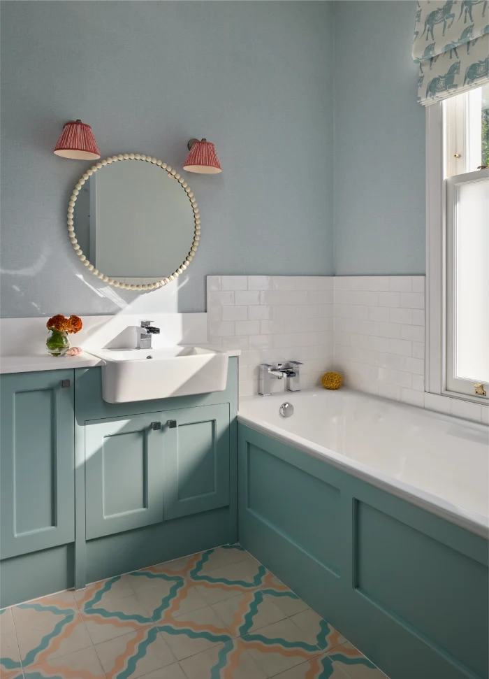 Charming bathroom with teal cabinetry, a round mirror, patterned floor tiles, and a subway-tiled bathtub.