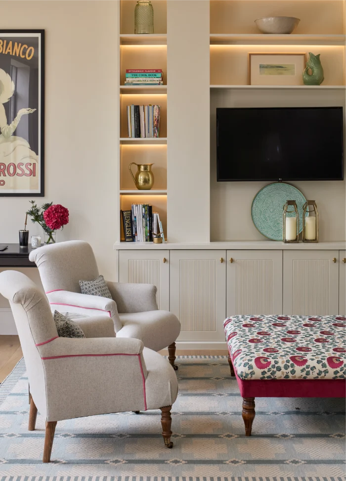 Elegant living room with built-in shelves, gray armchairs, a patterned ottoman, and warm accent lighting.