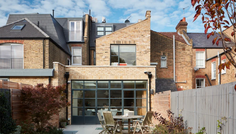 Brick townhouse exterior with a modern glass extension, outdoor dining area, and vibrant autumn foliage.