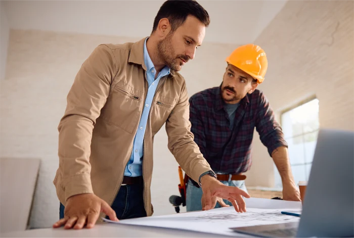 Two professionals reviewing blueprints at a construction site, with one wearing a hard hat and the other pointing at the plans.