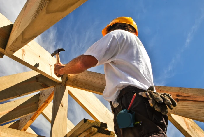 Construction worker wearing a yellow hard hat and gloves, hammering wooden beams under a bright blue sky.