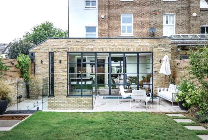 Modern brick house extension with large black-framed glass windows and doors, featuring a spacious patio area with outdoor seating, lush greenery, and a well-manicured lawn.