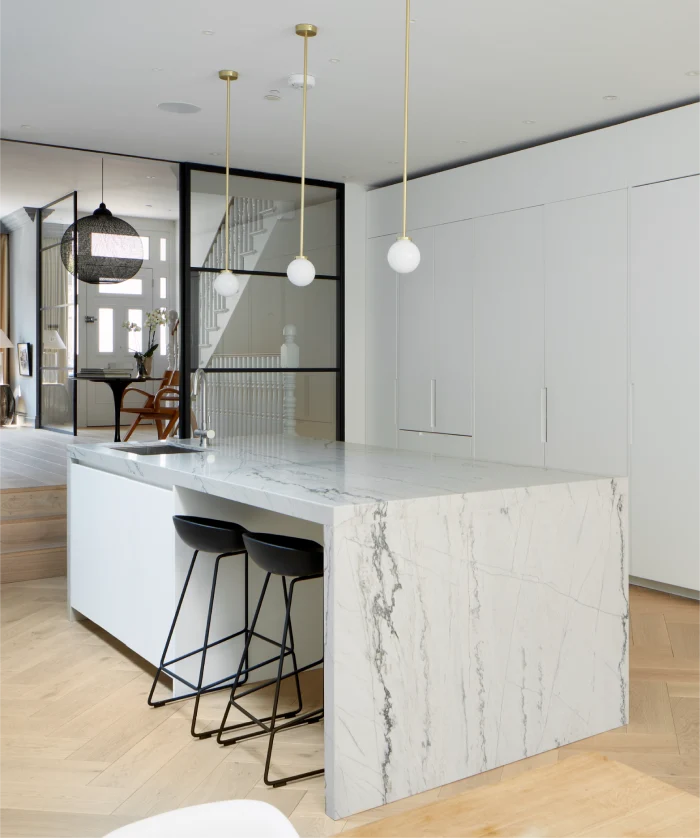 Modern kitchen interior featuring a marble island with bar stools, pendant lights, and glass partitions separating the staircase area.