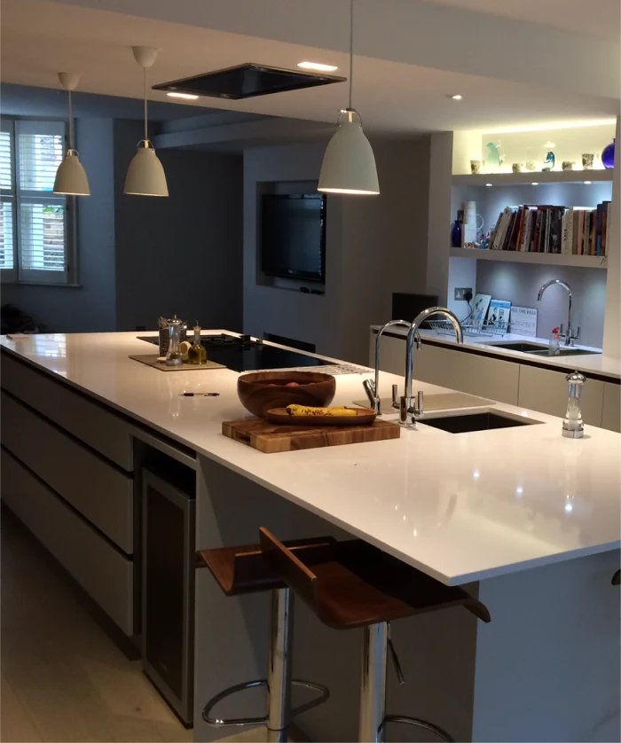 Modern kitchen with a sleek white island countertop, built-in appliances, pendant lighting, and wooden bar stools, featuring open shelving and ambient lighting.