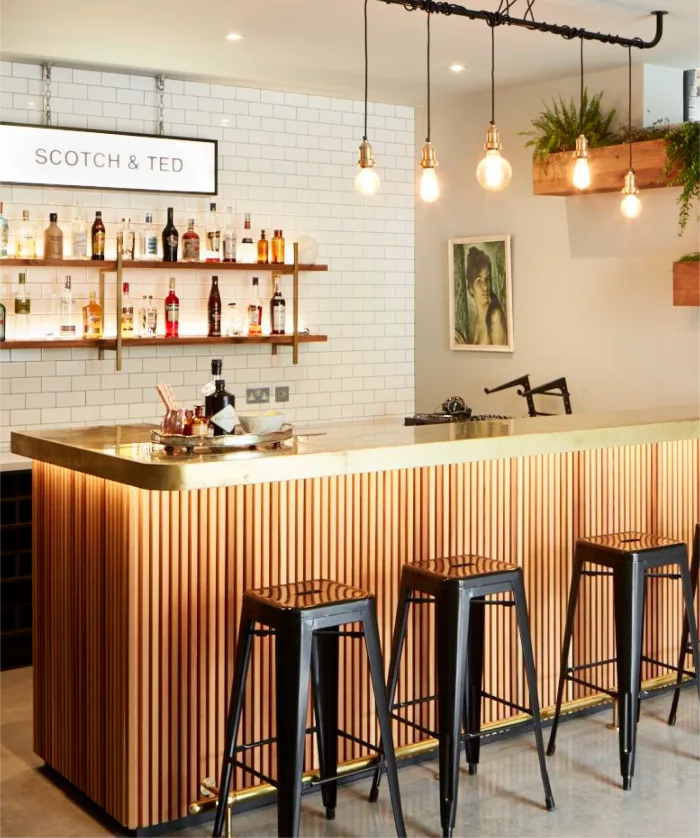 Modern bar with wooden slats, brass top, black stools, pendant lights, and white subway tile backdrop.