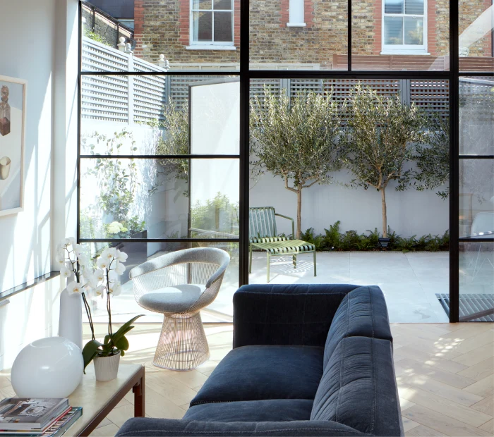 Bright and airy living space with a navy sofa, glass doors opening to a patio with olive trees, and modern decor.