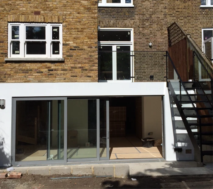 Modern brick house with large sliding glass doors on the ground floor, a balcony with a black railing on the first floor, and an external staircase leading to the upper level.