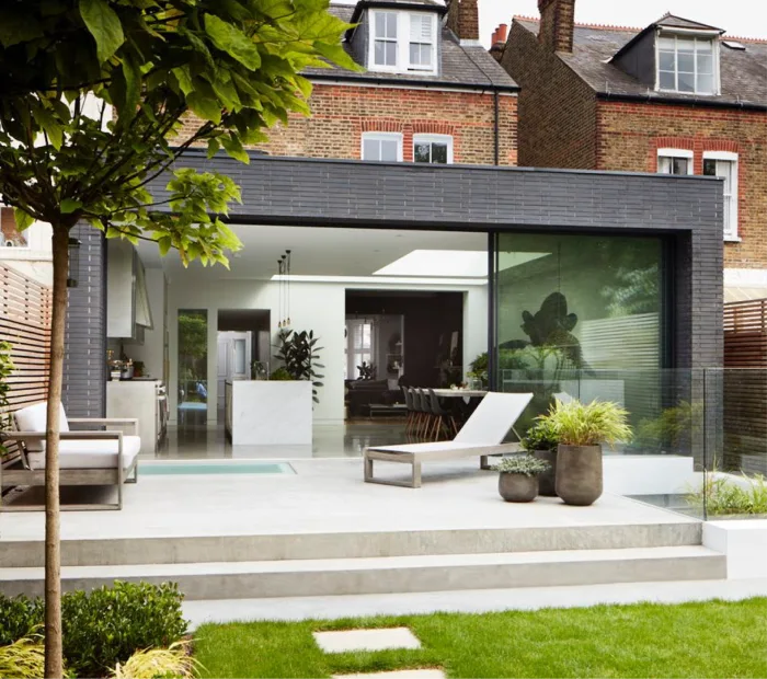 Contemporary backyard with open-plan extension, floor-to-ceiling glass doors, greenery, and modern lounge chairs.