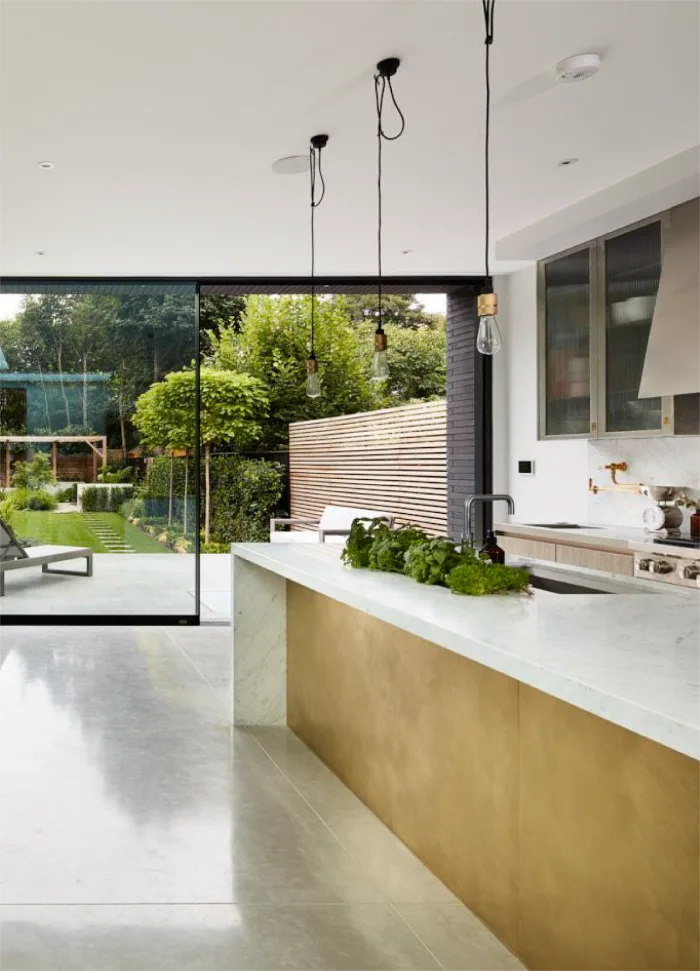 Modern kitchen with a sleek marble countertop, brass island panel, and large glass doors overlooking a lush backyard.