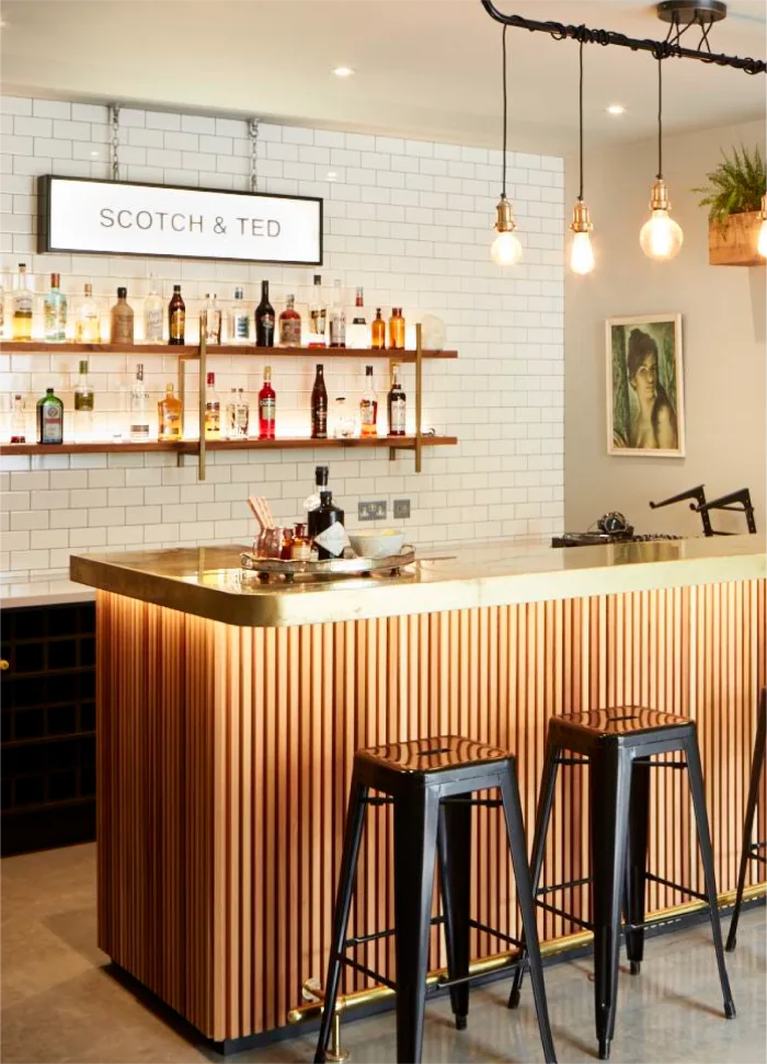 Modern home bar with wooden panel design, brass countertop, hanging lights, and shelves stocked with bottles.