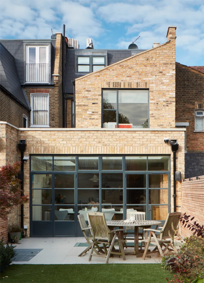 Charming brick exterior with glass doors leading to a patio featuring a wooden dining set and lush garden views.