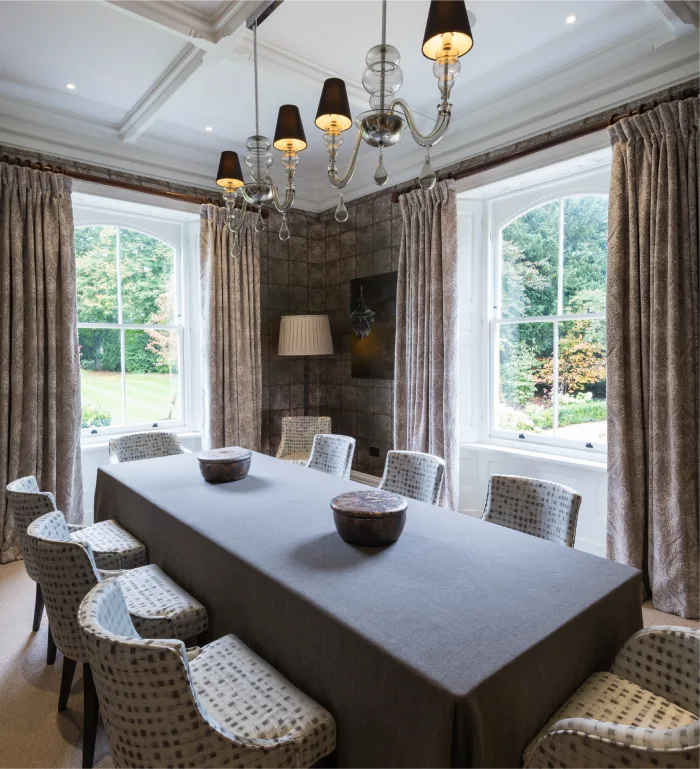 Elegant dining room with a long table, patterned chairs, chandeliers, and large windows overlooking a garden view.