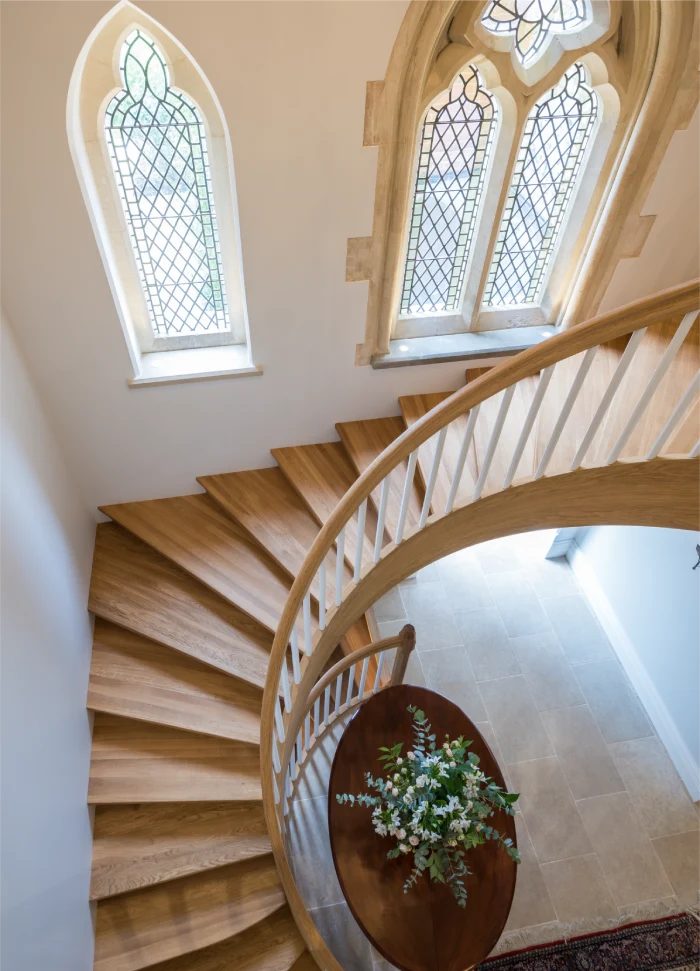 Curved wooden staircase with intricate banister and decorative windows, overlooking a floral arrangement.