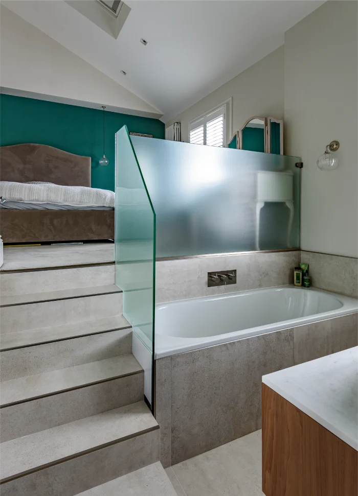 An innovative bedroom-bathroom combination, featuring a modern freestanding bathtub separated by frosted glass, with steps leading to a comfortable sleeping area accented by a bold teal wall.