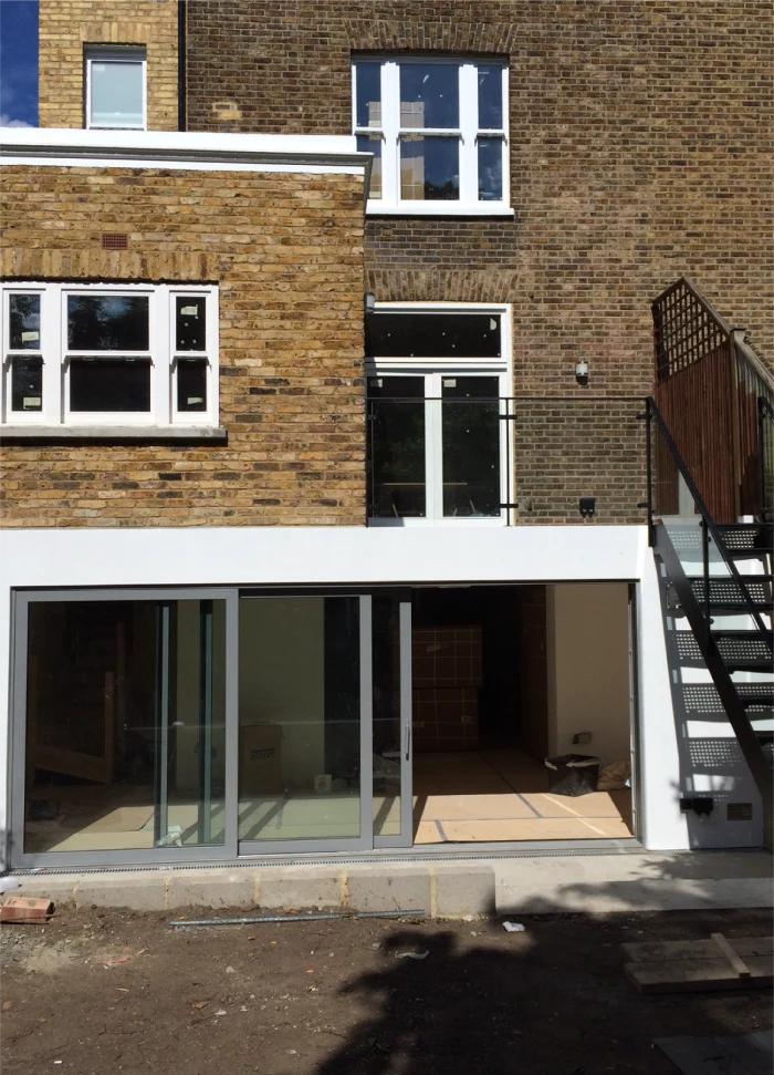 Exterior of a brick house with large white-framed windows, sliding glass doors on the ground floor, and a balcony with black metal railings. An outdoor staircase leads to the first floor.