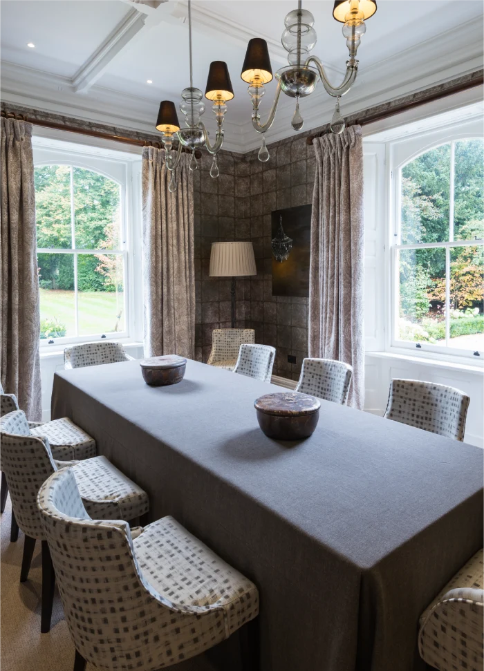 Formal dining room with a long table, patterned chairs, chandelier, and large windows overlooking greenery.