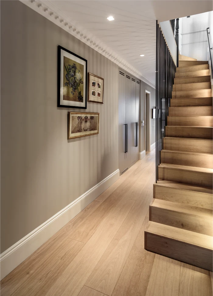 A sleek hallway design featuring wooden flooring, subtle wall paneling, and a modern staircase with built-in lighting and a black metal railing. The walls are adorned with framed artwork, adding a personal and elegant touch.