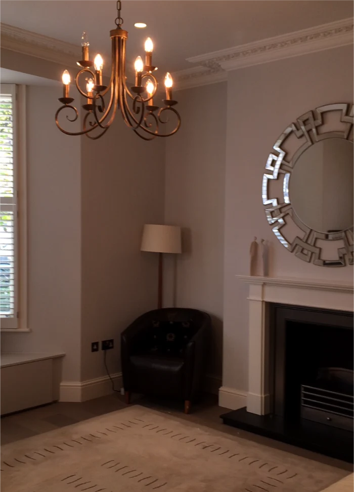 Living room with chandelier, ornate mirror, white mantel, black armchair, and natural light from windows.