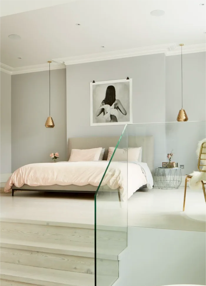 Minimalist bedroom with soft pink bedding, pendant lights, framed wall art, and sleek glass stair railing.