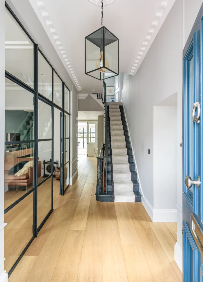 An inviting entryway with a contemporary design, showcasing natural wood flooring and a long hallway illuminated by a geometric pendant light. Black-framed glass partitions add a modern touch, providing glimpses into the adjacent spaces. A classic staircase with a neutral carpet runner leads upward, complemented by a vibrant blue door that adds character to the entrance.