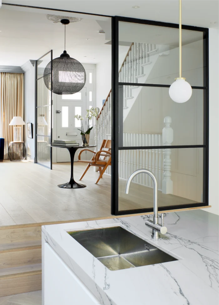 Modern kitchen sink with a marble countertop, featuring a view of a stylish open-plan living area with glass partitions and a staircase.