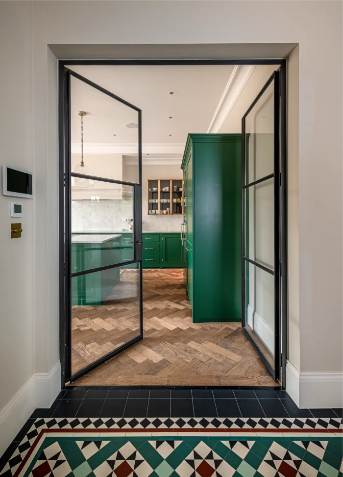 Stylish kitchen entrance with glass doors, herringbone wooden flooring, and bold green cabinetry visible through intricate patterned tiles.