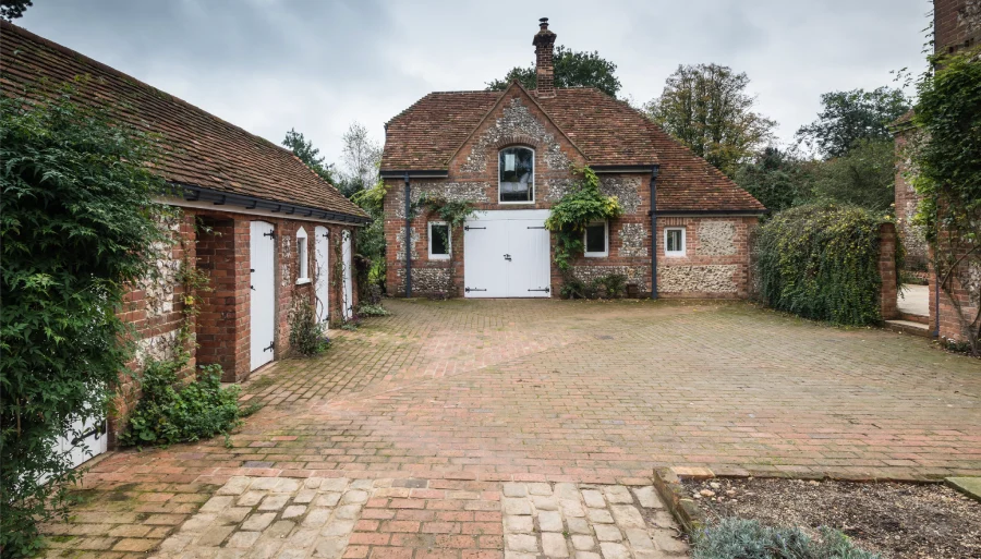 Rustic brick and flint outbuilding with white doors, surrounded by greenery and a paved courtyard.