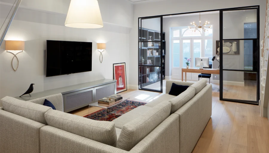 Contemporary living room with a sectional sofa, wall-mounted TV, and adjacent study area separated by glass sliding doors.