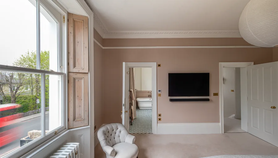 Elegant bedroom with a large window, vintage chair, wall-mounted TV, and a doorway view of a classic bathroom with a freestanding tub.