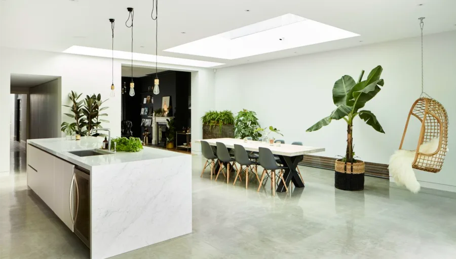 Modern open-plan kitchen and dining space featuring skylights, greenery, marble countertops, and a hanging chair.